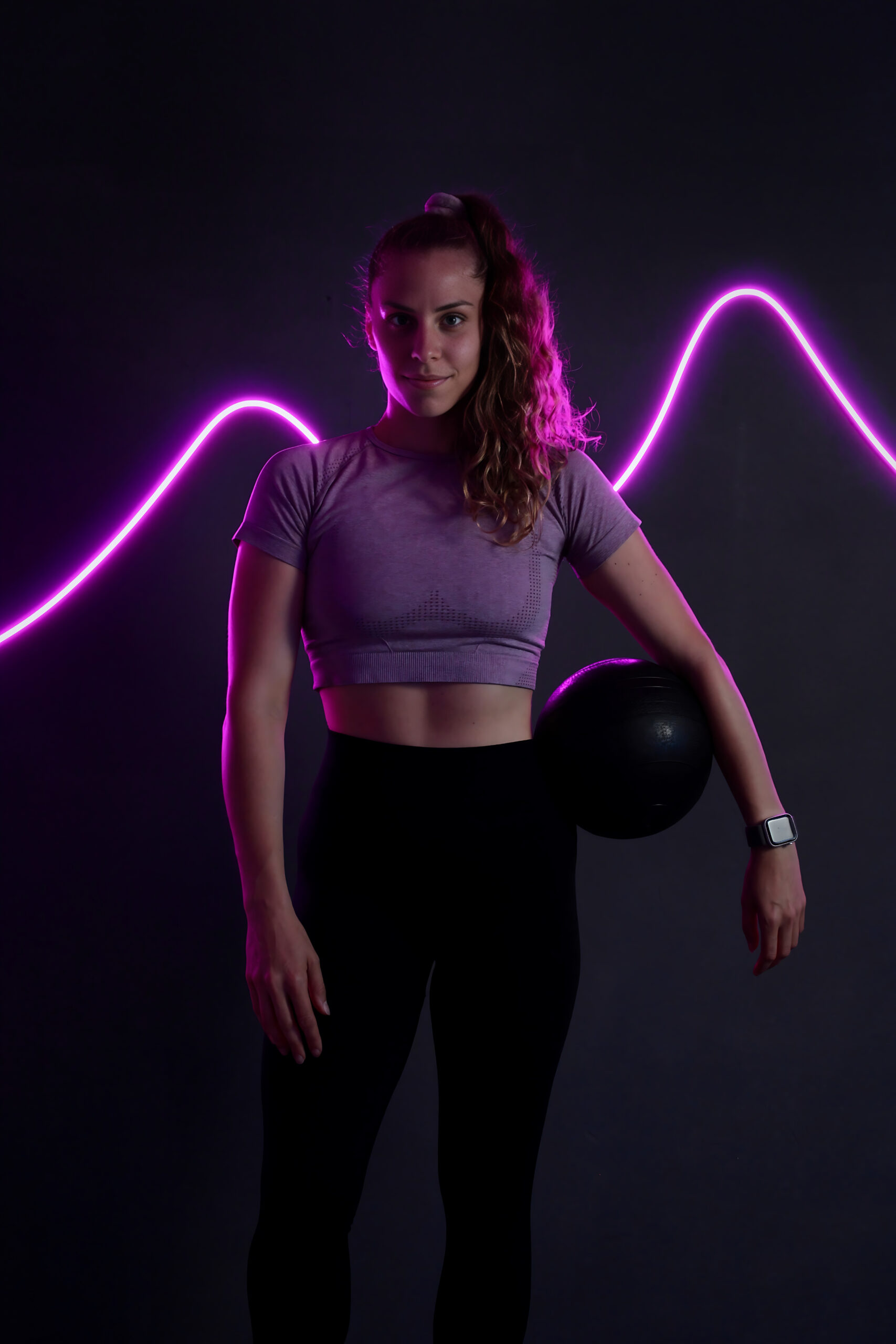 Tina Heller mit einem Medizinball vor der pinken Neonwelle im Studio